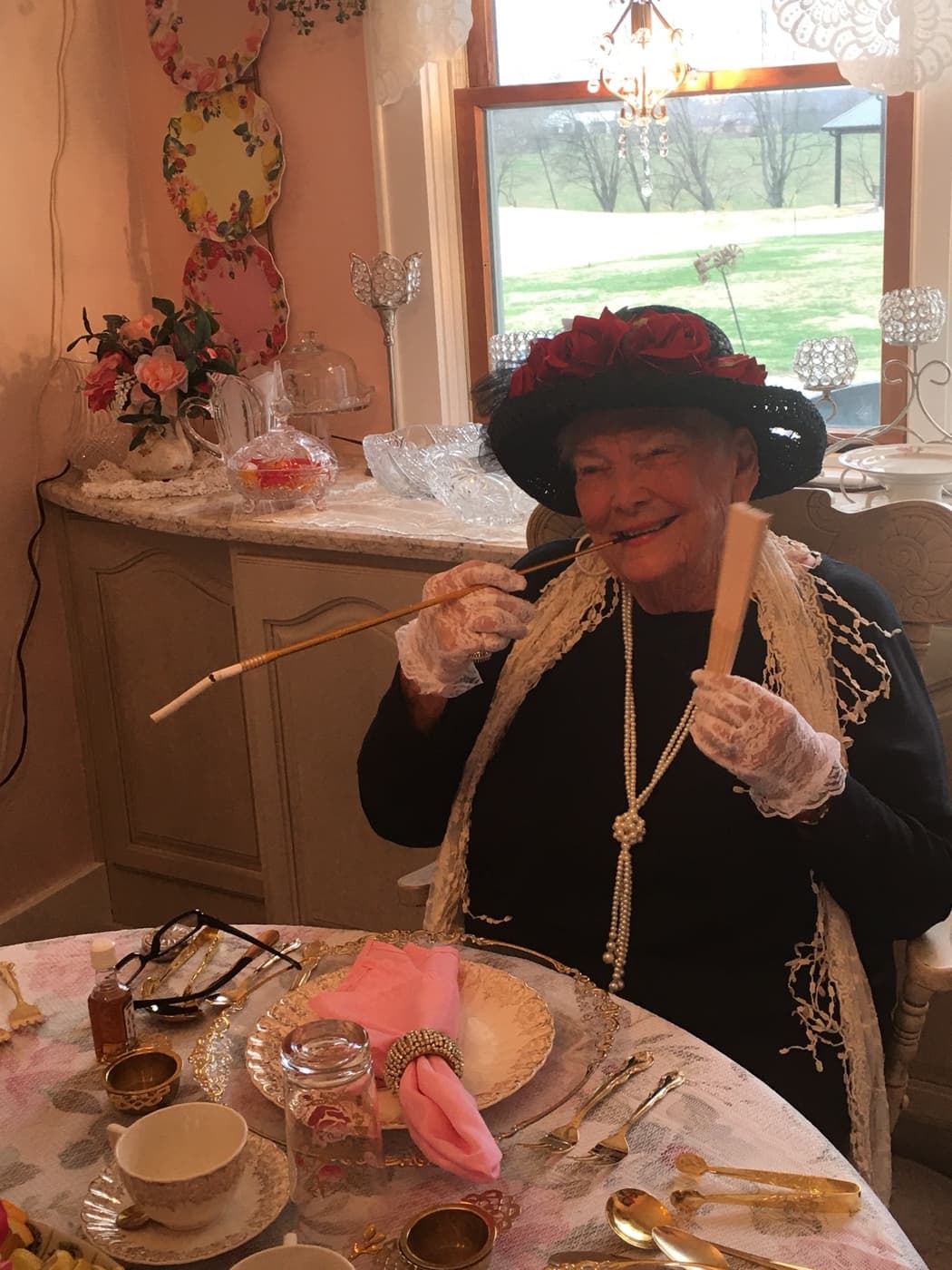 Ruth King in full Victorian costume, a hat banked with red roses, pearls, lace gloves and a long cigarette holder